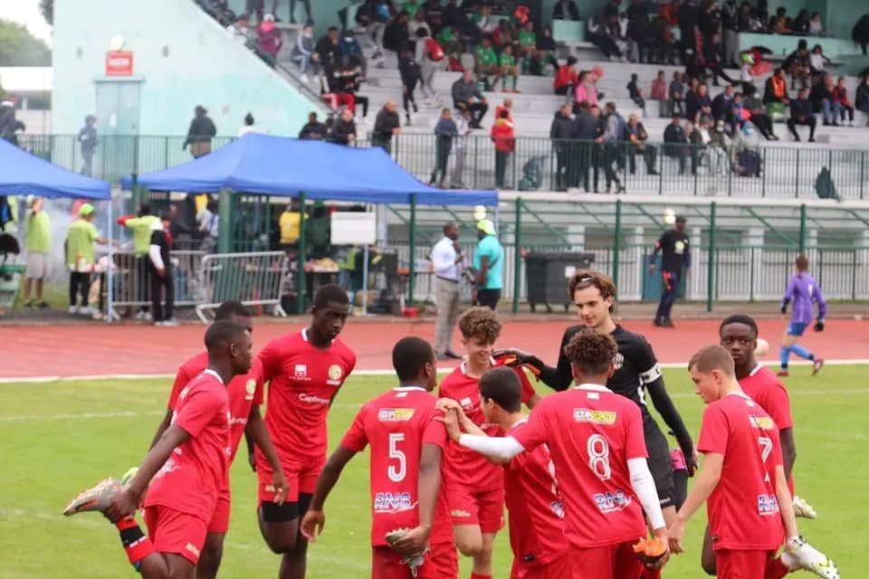 Une équipe de jeunes joueurs en maillot rouge se rassemble sur la pelouse, devant les stands de restauration du tournoi